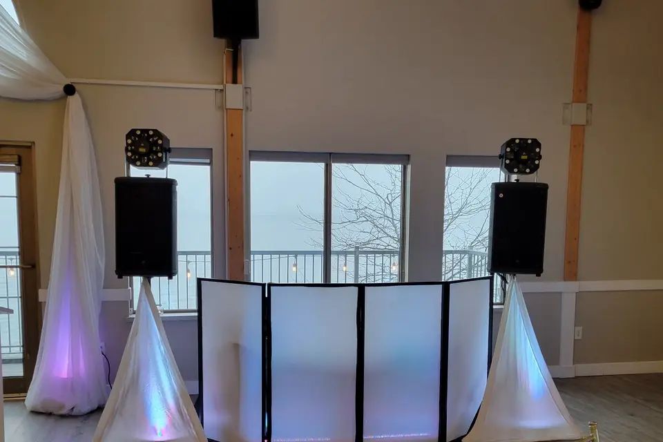 DJ booth, speakers and uplighting set up in a bright waterfront reception room